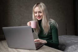 Smiling, joyful blonde girl in green golf works at home with a laptop with a cup of tea. Online video conference call. Nice talking. Online learning. Freelance.