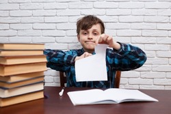 Stressed and exhausted schoolboy tearing papers sitting at the desk with blank note book. No ideas, studying difficulties, education, school concept
