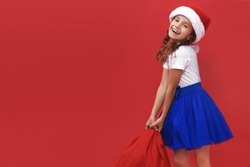Happy little smiling girl wearing santa hat carrying gift bag isolated on red background. Copy space. Horizontal view.