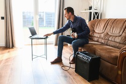 Online studying. A young man is watching video tutorial, video classes how to play guitar, he sits on couch with electric guitar and combo amp near
