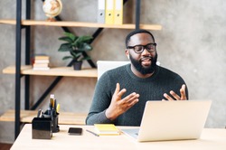 Video call. Attractive African-American guy is using laptop for video communication, a guy in glasses sits at the table in modern office, looking into webcam and talking online
