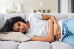 Sad young African American woman wearing casual clothes suffering from menstrual pain, feeling sick to her stomach, holding belly, having abdominal cramps during period and lying down on bed at home