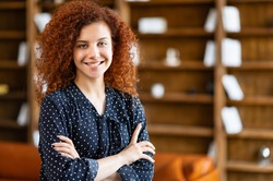 Ambitious smiling young curly woman standing in the office space with her arms crossed and looking at camera, charming red-haired female student in the library, succesful female entrepreneur indoors
