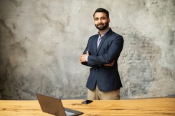 Indian businessman in formal suit stands in modern office near desk with a laptop and looking at camera. Ambitious smiling multiethnic man in smart casual wear with arms crossed on grey background