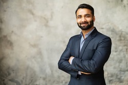 Smiling middle-age Indian high skilled leader in formal suit looks at the camera, confident ambitious bearded arabic businessman stands in office with arms folded on the grey background