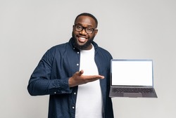 Smiling young African-American guy in casual wear showing laptop with empty display on grey background. Black guy points with palm at blank screen, advertising new app, presentation of newelty