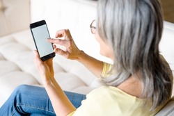 Back view a woman holding smartphone with an empty screen, senior gray-haired woman sits on couch and holds gadget with blank display, touching touchscreen with finger, mock-up, app advertising