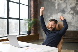 Excited young businessman in smart casual wear celebrating successful deal, looking at monitor raising fists in triumph, overjoyed male entrepreneur done project, screaming yes sitting at office