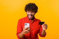 Cheerful middle eastern guy is using smartphone and credit card for shopping online. Happy man is ordering food online, male paying for long-awaited purchase on the mobile phone, isolated on orange