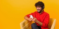 Studio portrait of young middle eastern guy in wireless headphones isolated on orange wall, looking at mobile phone, enjoying listening to favorite music soundtrack