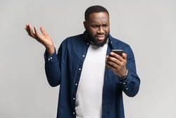 Disappointed young African-American man holding smartphone and watching at the phone screen, guy received bad notification or messages, feels upset, shrugging, isolated