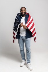 Portrait of positive african-american guy covered with usa flag, football lover support american nation league world cup, covered with flag wear denim jeans isolated over gray background, full length