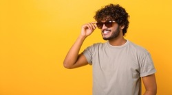 Stylish handsome Indian young curly man wearing casual t-shirt and sunglasses has good mood, enjoying sunny day, posing isolated on yellow background