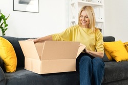 Happy senior mature adult woman is looking at her new parcels, sitting on the couch, and unpacking. Smiling lady opening online store order in the postal delivery shipping box