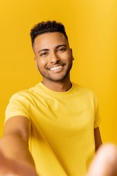 Nice attractive Indian man taking selfie, wearing casual t-shirt smiling joyfully looking at camera standing isolated on yellow background, pov arab man making video call, recording video