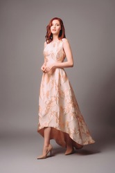 Ginger young woman in golden peach dress with floral print. Calm studio portrait of young lady in long sleeveless evening gown on grey background.