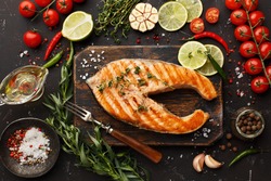Grilled salmon steak on vintage wooden board with fork, olive oil, vegetables, herbs and spices on dark concrete background. Top view