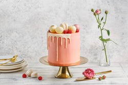 Tall pink cake decorated with macaroons, raspberries and chocolate balls on golden cake stand over white background with flowers and berries. Side view, copy space
