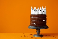 Chocolate Halloween cake with spooky meringue ghosts and candy eyes against an orange background with copyspace to side