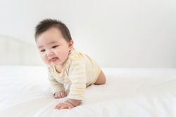 Portrait of cute little asian baby girl wearing bodysuit lying on white beedsheets at home. Cute infant child crawling on bed in the bedroom. Selective focus, free copy space. Childcare Concept.