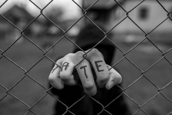 Aggressive teenage boy holding the wired fence at the correctional institute, the word hate is written on hes hand, conceptual image of juvenile delinquency in black and white.
