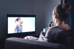 Woman watching romantic movie and eating ice cream. Sad lonely single girl streaming series or film on tv home at night. Person with stream on television. Comfy candid relaxing under blanket on couch.