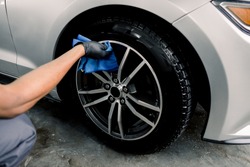 Car rims cleaning, car detailing wash concept. Cropped close up photo of male hand in black rubber glove with blue microfiber cloth washing car alloy wheel at car wash service