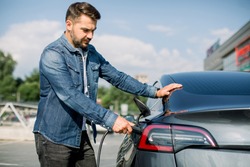 Side view of young handsome bearded man in jeans shirt, plugging wire into the car socket to charge his new luxury modern electric car at outdoor charging station in the city