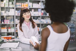 Positive young Caucasian woman pharmacist standing at the cashdesk in modern pharmacy, offering medication to African female customer. View through the shoulder of patient