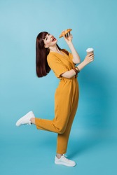 Food and drink promo ad. Full-length shot of young lady in elegant trendy ocher overalls and sneakers, posing on yellow background and holds delicious croissant and coffee to go
