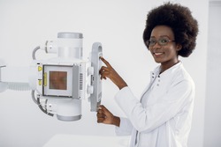 Beautiful African woman in white medical coat, standing near the X-Ray machine in modern hospital radiology room. X ray scanning of chest, heart, lungs in modern clinic