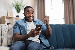 Surprised african guy sitting on couch and gesturing from happiness while reading good news on modern smartphone. Concept of people, success, technology and emotions.