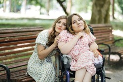 Portrait of two happy female friends looking at camera while spending free time at summer park. One woman sitting on wooden bench, another using wheelchair.