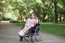 Portrait of pleasant young woman with spinal muscular atrophy smiling on camera among green summer park. Female person who using wheelchair. Concept of people with disability.