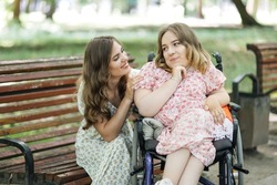 Portrait of two happy female friends looking at each other while spending free time at summer park. One woman sitting on wooden bench, another using wheelchair.