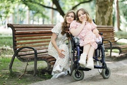 Two young ladies in beautiful summer dresses spending leisure time together at green city park. One woman sitting in wheelchair while another on wooden bench.