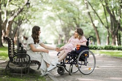 Smiling young woman sitting on wooden bench at summer park and holding hand of her female friend that using wheelchair. Real friendship with support. Leisure time outdoors.
