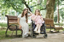 Two caucasian ladies in summer dressed smiling and looking at camera at green park. One woman sitting on wheelchair, another on wooden bench. Support for people with disability.