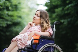 Female person with spinal muscular atrophy spending time outdoors alone with her thoughts. Young woman who using wheelchair sitting among green summer park and looking aside.