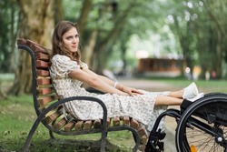Pleasant young woman keeping her feet on wheelchair while sitting on wooden bench among green summer park. Female person with disability spending time on fresh air.