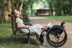 Side view of relaxed caucasian woman sitting on wooden bench at green park and holding legs on wheelchair. Casual lifestyles of person with disability.