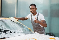 Young attractive African American man washing his yellow luxury car in a self-service car wash station showing thumb up outdoors with cleaning foam and yellow sponge.