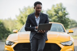 Handsome young African American business man working with laptop while standing near modern yellow cabrilet car outdoors, Handsome African man on background of sport car