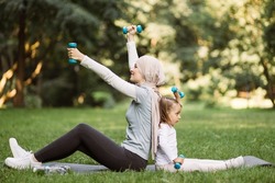 Fitness, workout, healthy living and gymnastics concept. Pretty Muslim mother and her cute daughter, sitting on the yoga mat and lifting dumbbells on fresh air at summer day