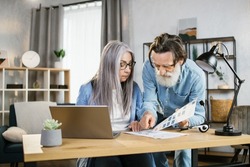Mature woman and bearded man business partners in casual wear discussing common project at bright office. Cheerful senior colleagues using modern gadgets for work.