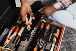 Close up of african american man in casual clothes taking pliers out of box at home. Focus on plastic black container with variety of modern tools for housework.
