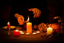Witch burns a herb on the altar in the dark. Female hands with sharp black nails do magic among candles, pumpkin, nuts, dry leaves, selected focus, low key.