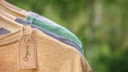 Organic clothes. Natural colored t-shirts hanging on wooden hangers in a row. Eco textile tag. Green forest, nature in background.