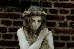 A young girl is dressed as a Halloween horror figure. 
She wears filthy rags as clothing and has a thorny 
crown with flowers on her head. She peers into the
camera with a sad expression on her face. 