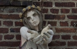 A young girl is dressed as a Halloween horror figure. 
She wears filthy rags as clothing and has a thorny 
crown with flowers on her head. She is seen begging 
with her hand reaching out.
